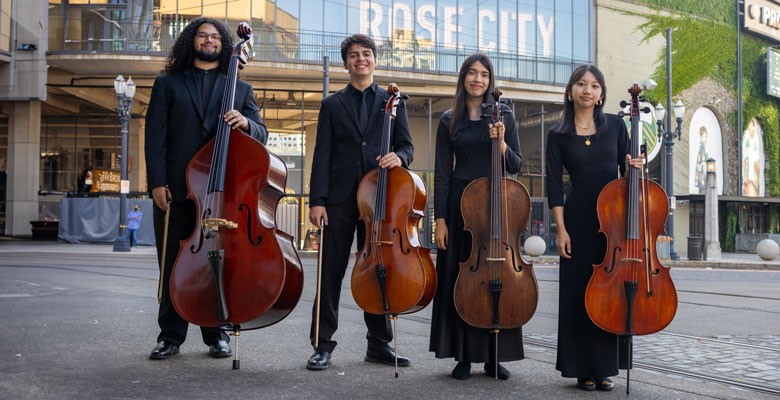 Four youth cello players stand on a street with their instruments