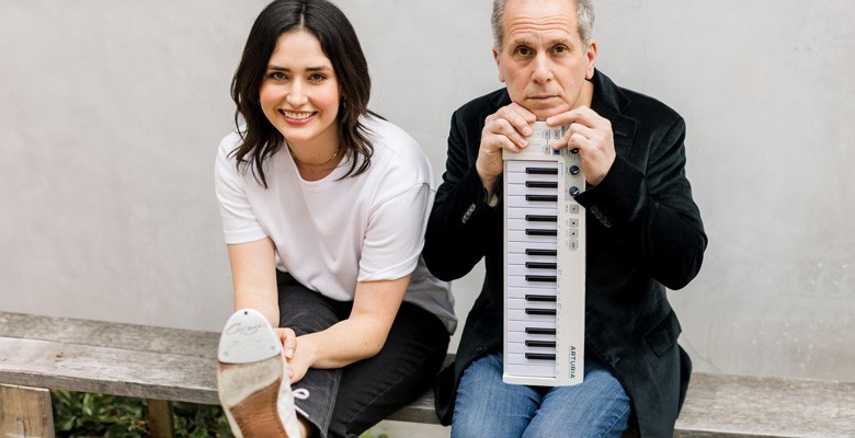 A photo of Larry Golding sitting with a small keyboard on the right and dancer Melinda Sullivan holding a foot up to the camera on the left