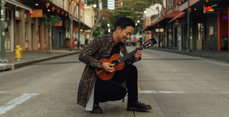 Jake Shimabukuro kneeling in the middle of an empty city street with his ukulele.