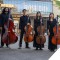 Four youth cello players stand on a street with their instruments